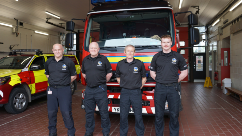 Four men in black t-shirts and trousers stand in a line in front of a fire engine.