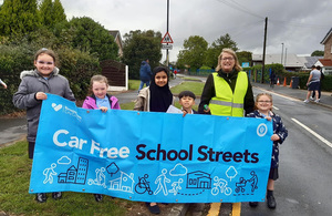 Credit: Active Travel England. A mixed group of children and adults stand together holding a banner that reads: 'Car free School Streets'.