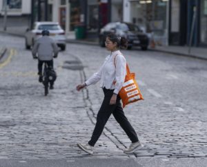 A woman is seen walking down a street with an orange living streets branded bag, reflecting a moment of everyday life and urban exploration.