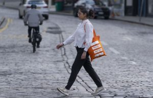 A woman is seen walking down a street with an orange living streets branded bag, reflecting a moment of everyday life and urban exploration.