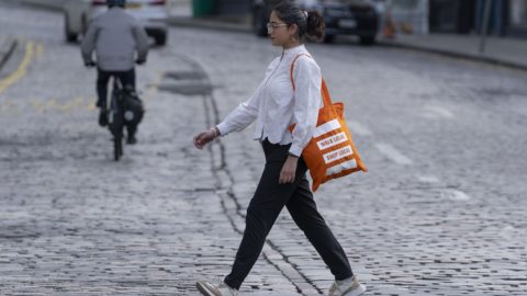 A woman is seen walking down a street with an orange living streets branded bag, reflecting a moment of everyday life and urban exploration.