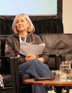 Woman sits on a stage, large screen in the background. Coffee table with microphone in front of her. She wears a Modeshift branded grey hoodie.