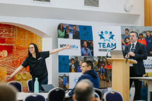 A man in a suit is speaking at a podium while a woman in a black "Modeshift" polo shirt excitedly gestures with open arms. Behind them, a large backdrop features the "Team Modeshift" logo and various images of people participating in sustainable travel initiatives. The audience is visible in the foreground, engaged in the presentation.