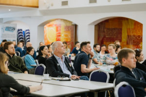 A diverse audience is seated at long tables in a conference room, attentively listening to a speaker. Some attendees are taking notes, while others are smiling or nodding. The room features bright artwork on the walls and banners promoting "Modeshift STARS."