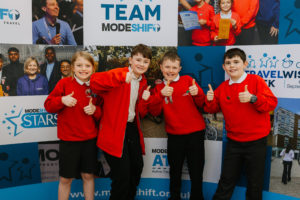 Four school children stand in a row. They are giving thumbs up gestures and smiling. Each wears a red school jumper. Behind them is a Modeshift branded display stand.