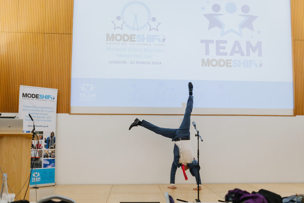 A performer in a pinstriped suit and red tie does a handstand on stage at a Modeshift STARS event. The backdrop features the 