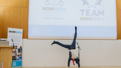 A performer in a pinstriped suit and red tie does a handstand on stage at a Modeshift STARS event. The backdrop features the "TEAM Modeshift" and "Modeshift STARS Education Masterclass Live" logos, along with the event details, "London | 20 March 2024." A Modeshift banner stands to the left, displaying the organisation's mission to support sustainable travel. A microphone stand is positioned on the stage.