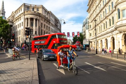 A street scene in London featuring a red double-decker bus, a black car, and a pedicab carrying two passengers with a cyclist pedaling in front. Union Jack flags hang above the street, and historic buildings line the background. Pedestrians walk along the pavement, enjoying the vibrant city atmosphere.