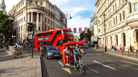 A street scene in London featuring a red double-decker bus, a black car, and a pedicab carrying two passengers with a cyclist pedaling in front. Union Jack flags hang above the street, and historic buildings line the background. Pedestrians walk along the pavement, enjoying the vibrant city atmosphere.