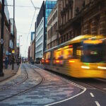 The image shows a vibrant urban street scene with a yellow tram in motion on tram tracks, surrounded by modern glass-fronted buildings and historical architecture. The street has a 30 mph speed limit sign, and a few pedestrians are visible walking along the sidewalks.