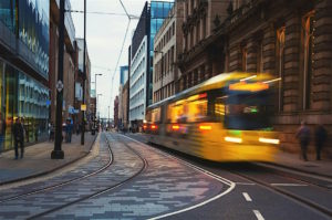  The image shows a vibrant urban street scene with a yellow tram in motion on tram tracks, surrounded by modern glass-fronted buildings and historical architecture. The street has a 30 mph speed limit sign, and a few pedestrians are visible walking along the sidewalks.