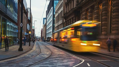 The image shows a vibrant urban street scene with a yellow tram in motion on tram tracks, surrounded by modern glass-fronted buildings and historical architecture. The street has a 30 mph speed limit sign, and a few pedestrians are visible walking along the sidewalks.