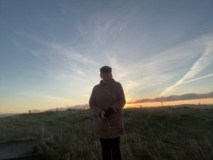 Woman stands smiling at camera. She wears a winter coat and hat. Behind here is a glowing sunrise and blue sky.