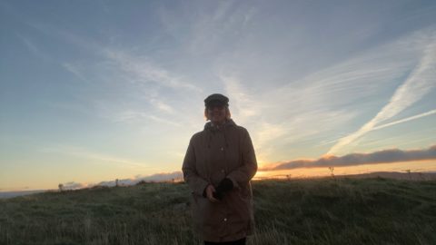 Woman stands smiling at camera. She wears a winter coat and hat. Behind here is a glowing sunrise and blue sky.