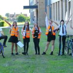 A group of seven students wearing school uniform stand in a row. The students on each end of the line hold bicycles. Four students in the center wear orange high viz vests. They all have their arms up in the air cheering. A school building a green playing field is in the background.