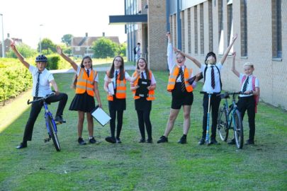 A group of seven students wearing school uniform stand in a row. The students on each end of the line hold bicycles. Four students in the center wear orange high viz vests. They all have their arms up in the air cheering. A school building a green playing field is in the background.