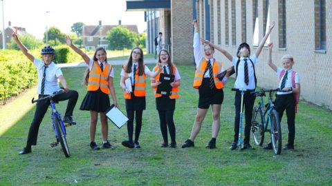 A group of seven students wearing school uniform stand in a row. The students on each end of the line hold bicycles. Four students in the center wear orange high viz vests. They all have their arms up in the air cheering. A school building a green playing field is in the background.