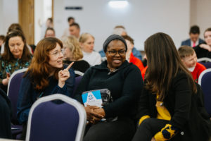 Audience at a conference., seated in rows of chairs. Three woman engaged in conversation. 