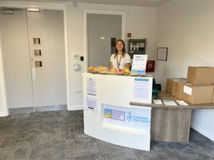 A woman with a friendly smile stands behind a reception desk at Colchester City Council. The desk has a "Colchester Borough Homes" and "Colchester City Council" logo on the front. There are fresh fruits, pastries, and an informational Modeshift STARS sign on the counter. Several cardboard boxes are stacked on a nearby table. The background features white walls, grey doors, and a fire safety panel.