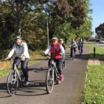 Image credit: Swindon Travel Choices. A group of older adults cycling along a dedicated bike path, surrounded by lush green trees. The cyclists, wearing helmets and casual attire, are smiling and riding in a relaxed manner, with a road and residential houses visible in the background.