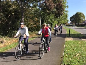 Image credit: Swindon Travel Choices. A group of older adults cycling along a dedicated bike path, surrounded by lush green trees. The cyclists, wearing helmets and casual attire, are smiling and riding in a relaxed manner, with a road and residential houses visible in the background.