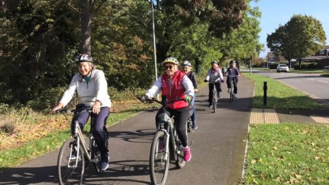 Image credit: Swindon Travel Choices. A group of older adults cycling along a dedicated bike path, surrounded by lush green trees. The cyclists, wearing helmets and casual attire, are smiling and riding in a relaxed manner, with a road and residential houses visible in the background.