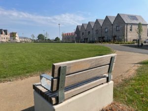 Image credit: Swindon Travel Choices. A peaceful residential area with a large green open space and a wooden bench in the foreground. A row of modern brick houses with pitched roofs and solar panels lines the background, while a paved footpath curves around the park under a clear blue sky.