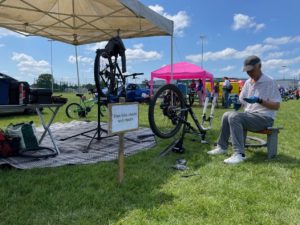 Image credit: Swindon Travel Choices. A community event featuring a free bike repair station in a grassy field. Under a canopy, a mechanic is working on a bicycle mounted on a stand, while another person sits on a bench handling tools. A sign reading "Free bike checks and repairs" is visible in the foreground, with families and event tents in the background.