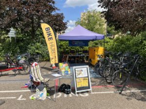 An outdoor cycling event setup with a "BIKE HUB" banner and a "Colchester Bike Kitchen" tent. Several bicycles are parked, and a yellow cargo bike is visible. A small table with informational leaflets and a sign advertising a cycling event is placed in front of the tent. The surrounding area includes trees, bushes, and a brick building under a blue sky.