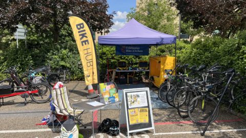 An outdoor cycling event setup with a "BIKE HUB" banner and a "Colchester Bike Kitchen" tent. Several bicycles are parked, and a yellow cargo bike is visible. A small table with informational leaflets and a sign advertising a cycling event is placed in front of the tent. The surrounding area includes trees, bushes, and a brick building under a blue sky.