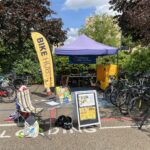 An outdoor cycling event setup with a "BIKE HUB" banner and a "Colchester Bike Kitchen" tent. Several bicycles are parked, and a yellow cargo bike is visible. A small table with informational leaflets and a sign advertising a cycling event is placed in front of the tent. The surrounding area includes trees, bushes, and a brick building under a blue sky.