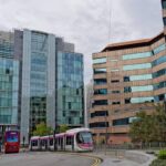 A modern tram with pink and white accents travels through an urban setting in Birmingham, UK, alongside a red double-decker bus. The scene is framed by contemporary glass and steel buildings on the left and a distinctive brown, angular office building with reflective windows on the right. The sky is overcast, and greenery lines the streets.