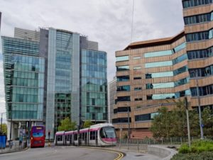 A modern tram with pink and white accents travels through an urban setting in Birmingham, UK, alongside a red double-decker bus. The scene is framed by contemporary glass and steel buildings on the left and a distinctive brown, angular office building with reflective windows on the right. The sky is overcast, and greenery lines the streets.