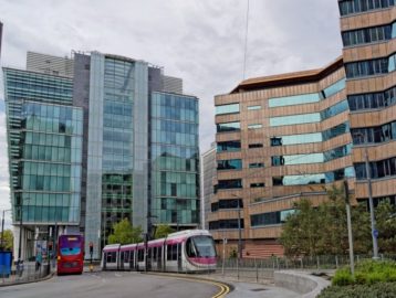 A modern tram with pink and white accents travels through an urban setting in Birmingham, UK, alongside a red double-decker bus. The scene is framed by contemporary glass and steel buildings on the left and a distinctive brown, angular office building with reflective windows on the right. The sky is overcast, and greenery lines the streets.