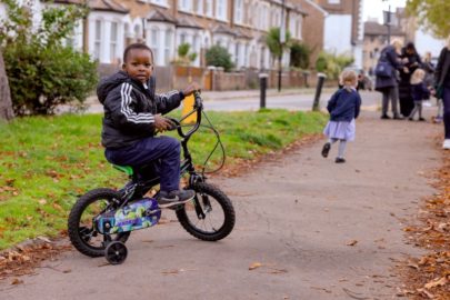 Image credit: Active Travel England. A young boy wearing a black jacket sits on a small black and green bicycle with training wheels on a paved path. He looks back towards the camera with a curious expression. In the background, a young girl in a navy blue top and light blue dress walks away, and a group of people is gathered further down the path. The setting is a residential street with a row of houses and greenery lining the pavement.