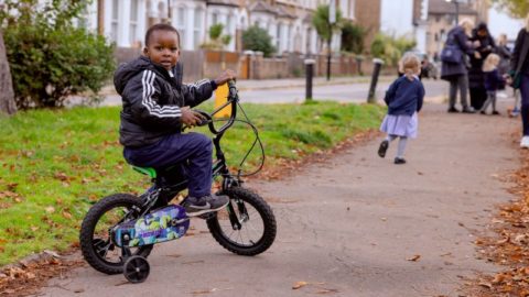 Image credit: Active Travel England. A young boy wearing a black jacket sits on a small black and green bicycle with training wheels on a paved path. He looks back towards the camera with a curious expression. In the background, a young girl in a navy blue top and light blue dress walks away, and a group of people is gathered further down the path. The setting is a residential street with a row of houses and greenery lining the pavement.