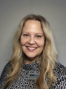 A woman with long blonde hair smiles at the camera. She is wearing a patterned blouse with a high neckline and a name badge that reads "MODESHIFT HONORARY MEMBER." The background is a neutral gray color.