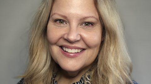 A woman with long blonde hair smiles at the camera. She is wearing a patterned blouse with a high neckline and a name badge that reads "MODESHIFT HONORARY MEMBER." The background is a neutral gray color.