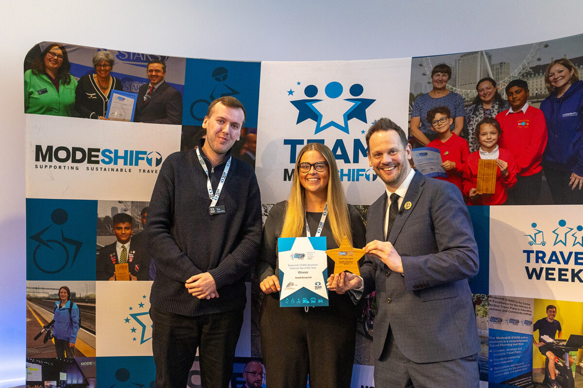A woman and two men smiling posing with wooden star shaped trophy and certificate. Modeshift branded display stand behind them.