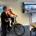 A woman sits on a stationary bike wearing a VR headset while a trainer stands beside her. A large screen displays a virtual cycling environment showing a residential street with a yellow car. A Modeshift Active Travel Ambassador (ATA) Program banner is visible in the background.