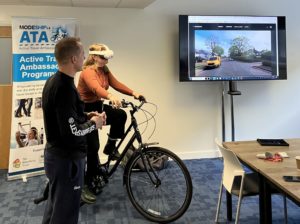  A woman sits on a stationary bike wearing a VR headset while a trainer stands beside her. A large screen displays a virtual cycling environment showing a residential street with a yellow car. A Modeshift Active Travel Ambassador (ATA) Program banner is visible in the background.