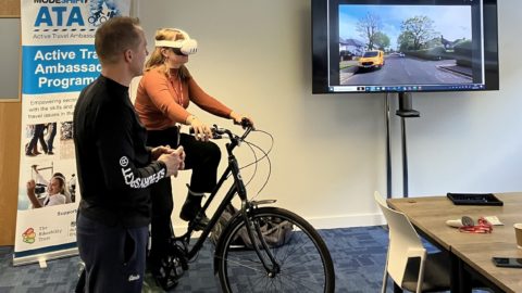 A woman sits on a stationary bike wearing a VR headset while a trainer stands beside her. A large screen displays a virtual cycling environment showing a residential street with a yellow car. A Modeshift Active Travel Ambassador (ATA) Program banner is visible in the background.