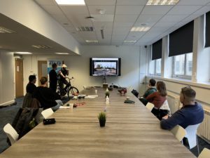  A group of people sits around a large meeting table watching a VR cycling training session. One participant is on a stationary bike wearing a VR headset, while a trainer provides guidance. A large screen shows a virtual cycling route, and the Modeshift ATA Program banner is displayed nearby.