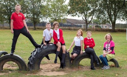 Group of six children and one adult sitting and posing on large, half-buried tires in a grassy outdoor playground. The children are wearing school uniforms, with red and white tops, while the adult wears a pink and black outfit. Trees and school buildings are visible in the background. Image credit: Living Streets