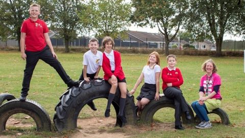 Group of six children and one adult sitting and posing on large, half-buried tires in a grassy outdoor playground. The children are wearing school uniforms, with red and white tops, while the adult wears a pink and black outfit. Trees and school buildings are visible in the background. Image credit: Living Streets