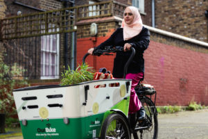 A woman wearing a pink headscarf rides a cargo bike past a brick building. Showcasing a blend of mobility and practicality.