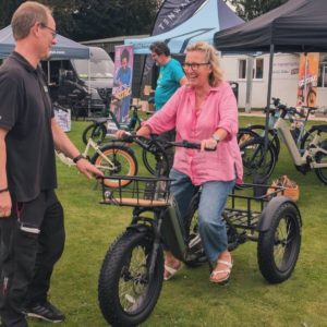 She's Electric Local event in Aylsham. A woman wearing a pink shirt and jeans is smiling while sitting on an electric tricycle at an outdoor event. She is engaged in conversation with a man in black clothing who appears to be demonstrating the bike. The background features exhibition tents, banners, and various e-bikes on display, creating a lively and engaging atmosphere.