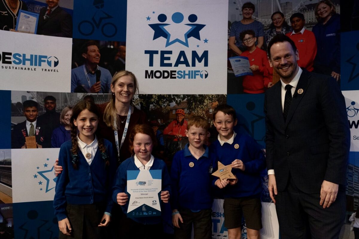 A group of four school children and two adults smile at the camera. Two of the children dressed in school uniform hold up a star shaped award and certificate.