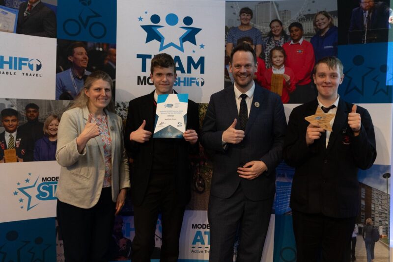 Four people smile, making thumbs up gestures. Two students in school uniform hold up a star shaped award and certificate.