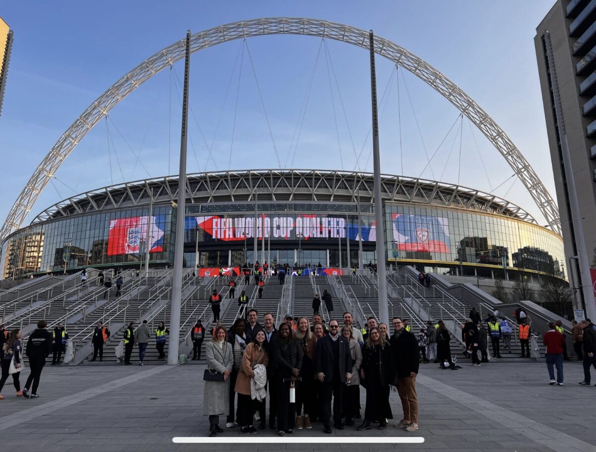 A group of people standing in front of Wembley Stadium, with the iconic arch visible in the background. The stadium is hosting a FIFA World Cup qualifier, as indicated by the signage, and security personnel and attendees are seen in the background near the entrance.